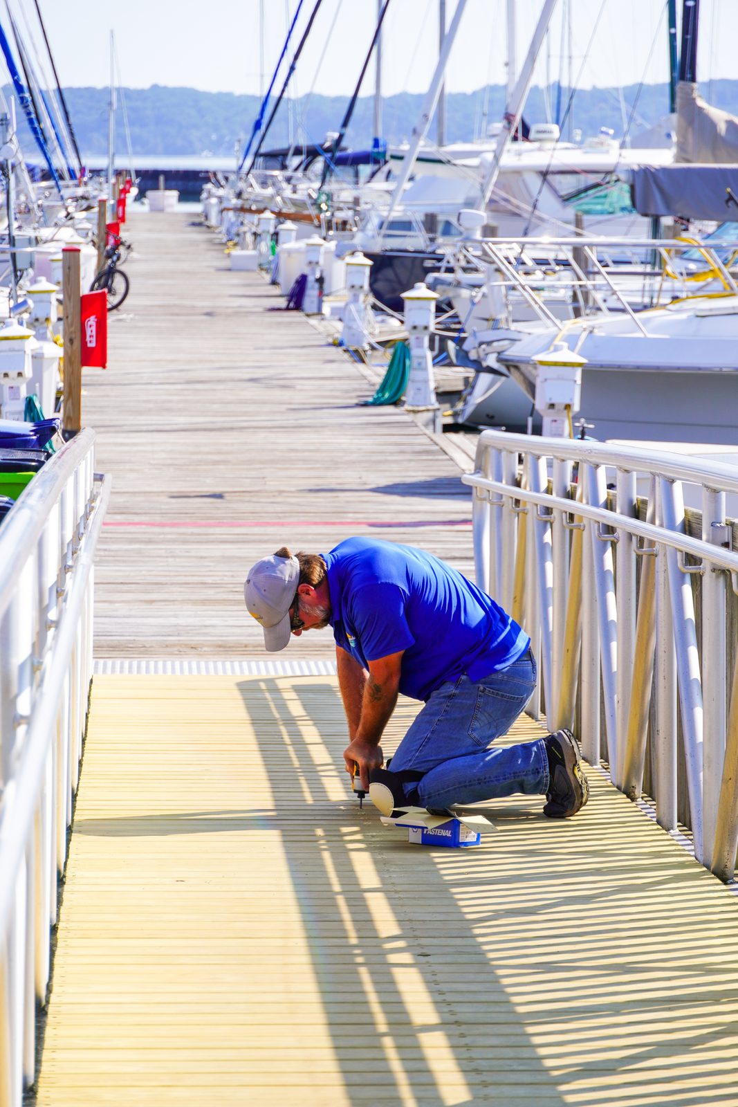 A dockhand kneeling to make a repair along the main dock, sailboats lining both sides.