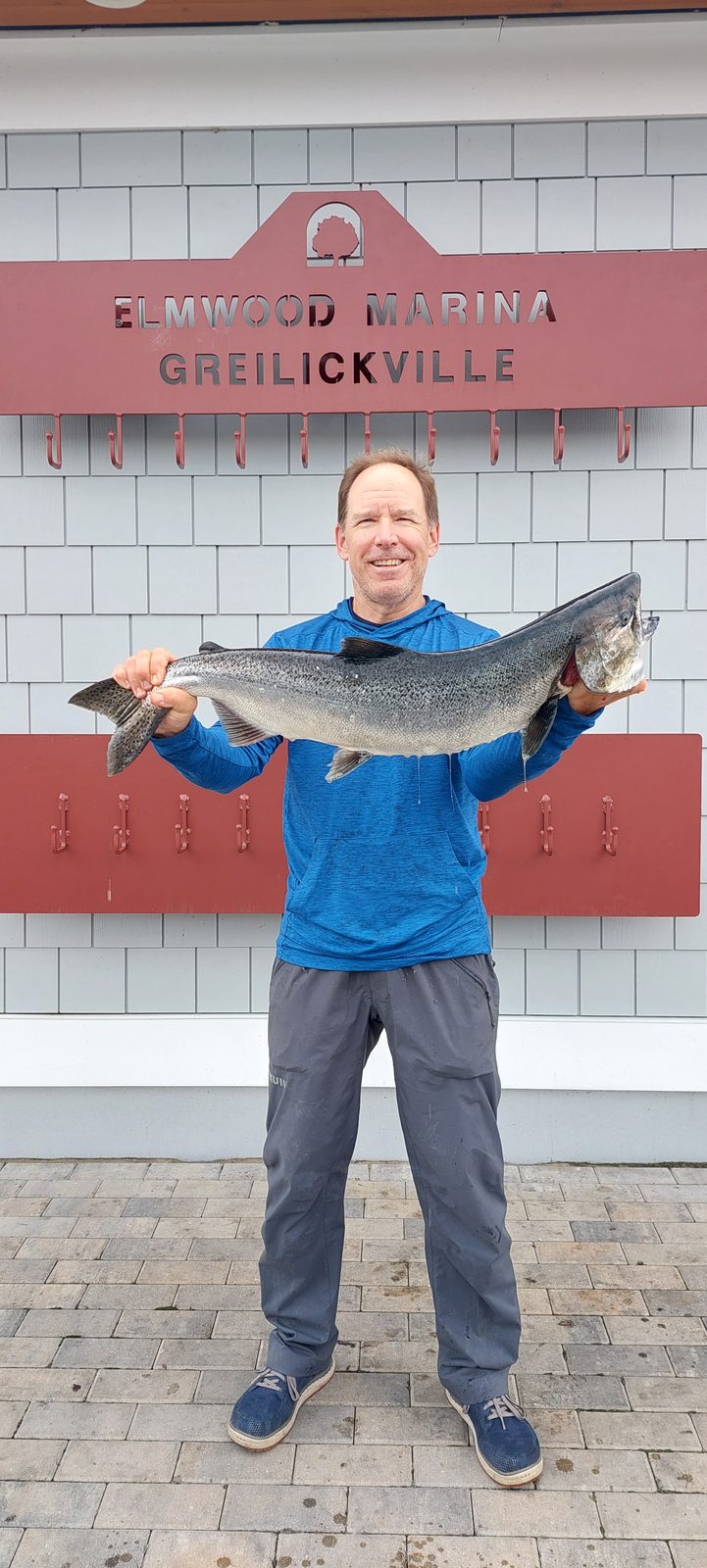 An angler proudly holding a large salmon in front of the Elmwood Marina sign.