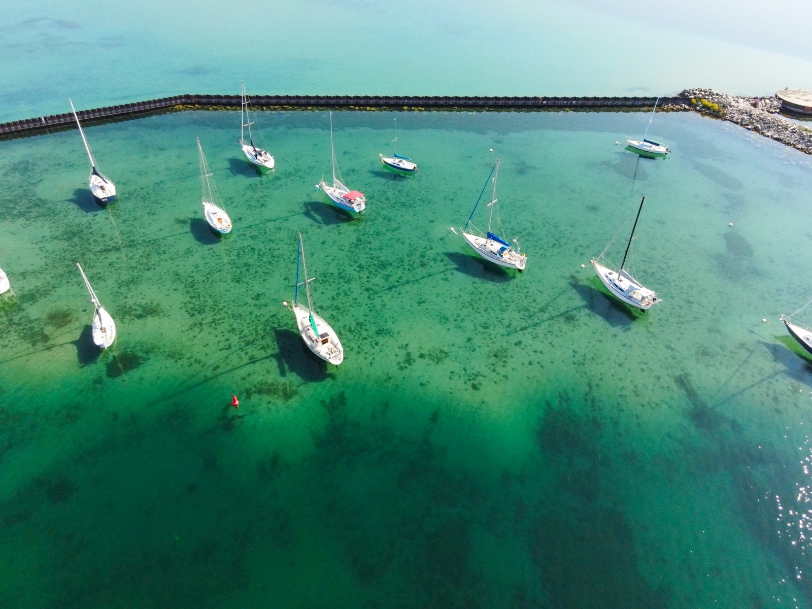 Aerial of sailboats riding on moorings in clear turquoise water inside the breakwall.
